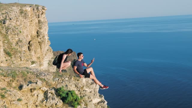 Young-backpacker-woman-approaching-man-sitting-on-edge-of-rock-couple-taking-selfie