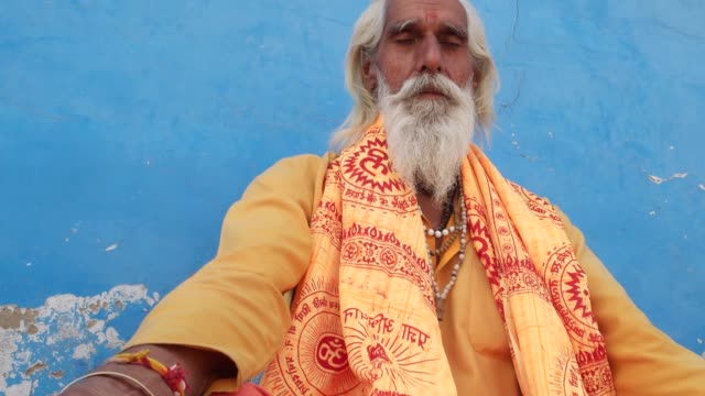 Old-Indian-sadhu,-saint,-sitting-and-meditation-with-closed-eyes-near-a-temple-in-Pushkar,-Rajasthan