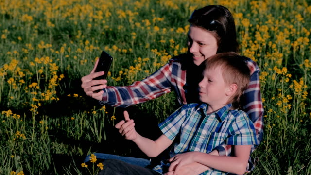Mutter-und-Sohn-machen-Selfie-am-Telefon-sitzen-auf-dem-Rasen-unter-den-gelben-Blumen.