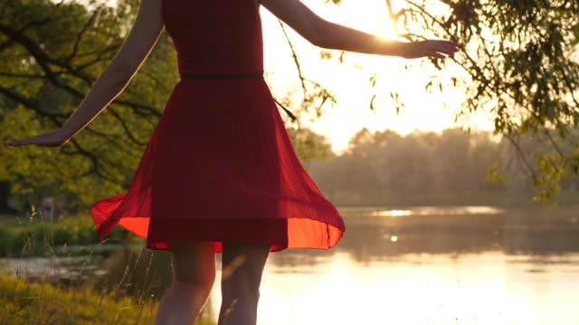 Woman-in-red-transparent-dress-dancing-and-spinning-around-against-lake-at-sunset