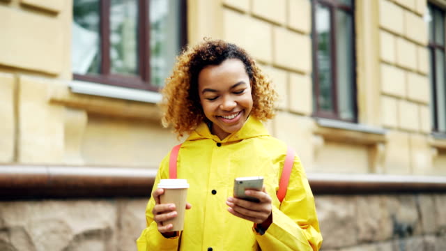 Mujer-de-raza-mixta-atractiva-está-utilizando-smartphone-moderno-mirando-la-pantalla-y-sonriendo-navegando-por-la-red-o-viendo-fotos-y-holding-tome-café-a-pie-al-aire-libre.