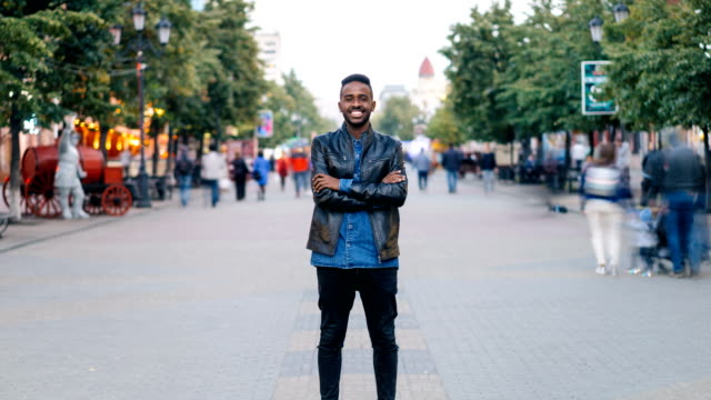 Zoom-in-time-lapse-of-happy-African-American-guy-wearing-jeans-and-leather-jacket-standing-alone-in-street-downtown,-smiling-and-looking-at-camera-with-crowd-moving-by.