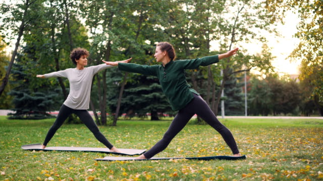 Chica-delgada-experimentado-instructor-enseña-a-su-estudiante-hatha-yoga-asanas-cálido-día-de-otoño-en-el-parque-de-la-ciudad.-Estilo-de-vida-saludable-y-deporte-para-el-concepto-de-zonas-urbanas.
