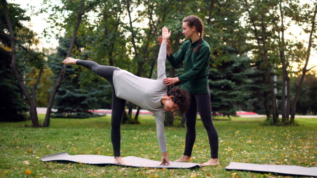 Friendly-yoga-instructor-is-helping-female-student-to-maintain-Half-Moon-pose-Ardha-Chandrasana-standing-near-her,-speaking-and-holding-her-hand-and-leg.