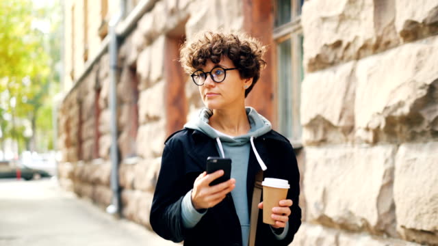 Beautiful-girl-with-dark-curly-hair-is-using-smartphone-texting-friends-and-holding-coffee-walking-in-city-alone.-Modern-technology,-communication-and-drinks-concept.