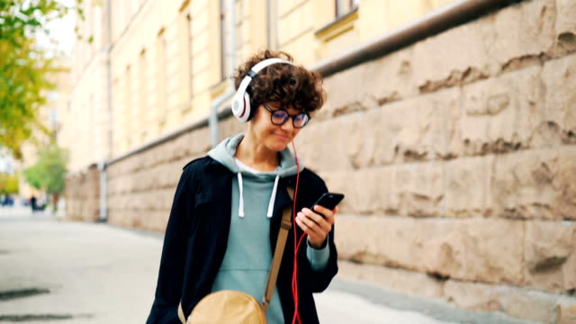 Fröhliche-junge-Dame-ist-Musik-über-Kopfhörer-hören-und-Smartphone-auf-Straße-Herbsttag-alleine-halten.-Kultur-und-Technik-Jugendkonzept.