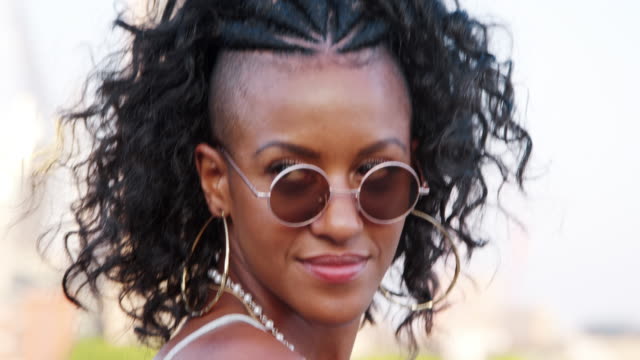 Trendy-young-black-woman-in-camisole-and-sunglasses-walking-over-bridge,-looking-back-and-laughing-to-camera,-close-up,-follow-shot