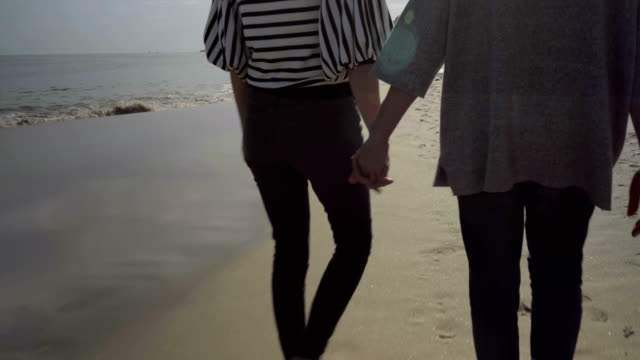 Two-women-holding-hands-together-while-walking-on-sandy-beach.