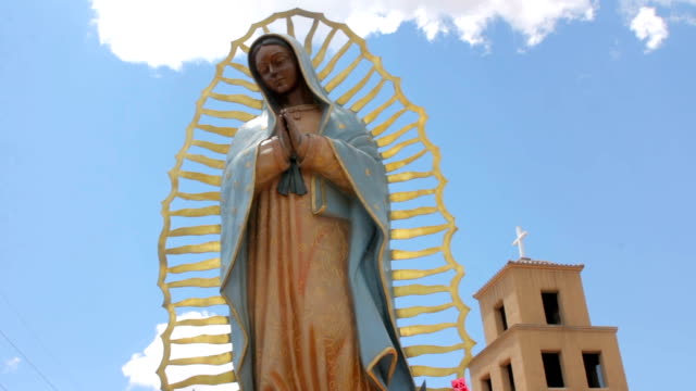 Ángulo-bajo-de-una-estatua-de-la-Virgen-Guadalupe-con-una-iglesia-de-Adobe