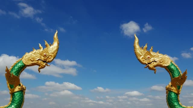 Dragon-statues-facing-each-other-at-Nakhon-Phanom,-Thailand.-Naga,-symbol-of-protection.-Time-lapse,-moving-clouds-against-clear-blue-sky.