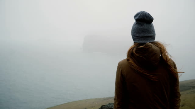 Back-view-of-young-woman-in-hat-standing-on-the-shore-of-the-sea-and-thinking-about-something,-exploring-the-Iceland