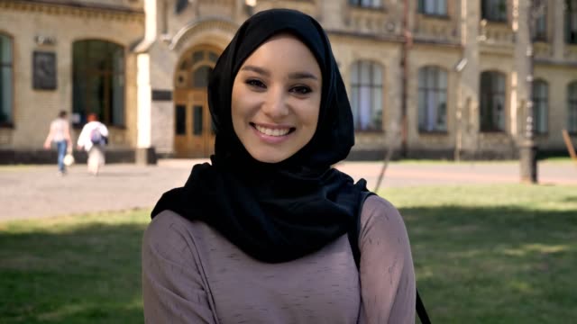 Junge-süße-muslimisches-Mädchen-Hijab-ist-stehend-und-lächelnd-in-tagsüber-im-Sommer,-gerade-in-die-Kamera,-aufbauend-auf-Hintergrund,-Religiuos-Konzept,-emotionales-Konzept
