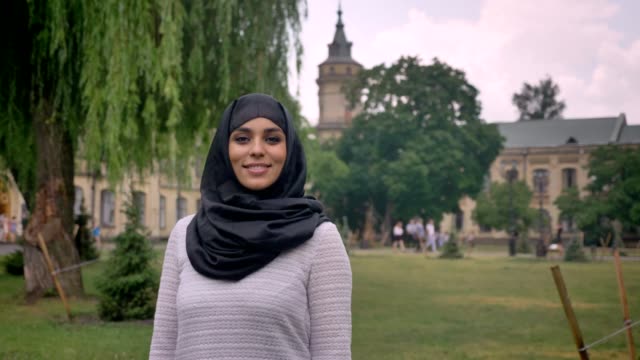 Junge-süße-muslimisches-Mädchen-Hijab-ist-stehend-und-lächelnd-in-tagsüber-bei-schlechtem-Wetter,-gerade-in-die-Kamera,-aufbauend-auf-Hintergrund,-Religiuos-Konzept