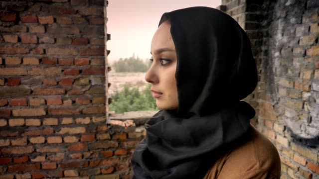 Joven-mujer-musulmana-triste-en-hijab-en-edificios-abandonados,-dando-vuelta-y-mirando-a-cámara,-concepto-de-miedo-y-terror,-terrorismo