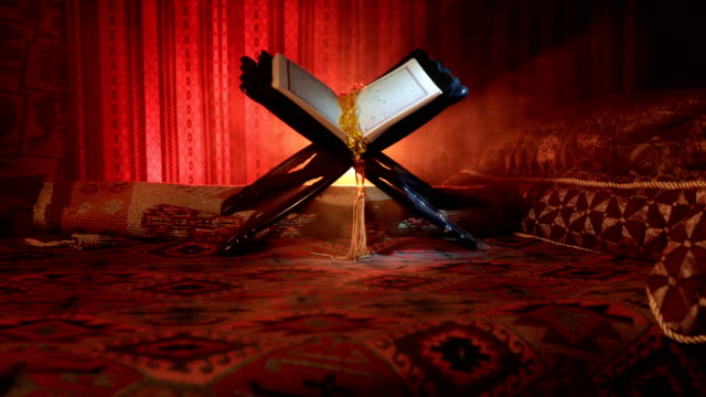 Open-holy-book-of-Muslims-on-stand-on-eastern-carpet-with-dark-toned-foggy-background.-Muslim-religion-concept.-Selective-focus.-Slider-shot.