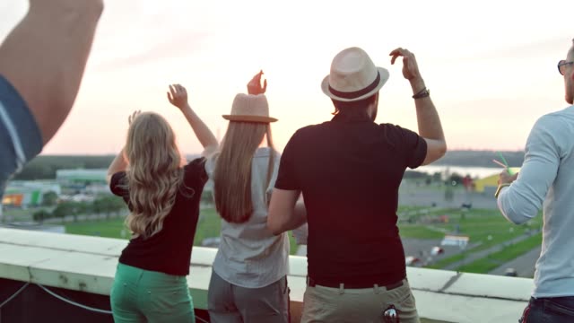 Rear-view-group-of-people-enjoying-beautiful-city-landscape-raising-arms-on-rooftop-party-at-sunset