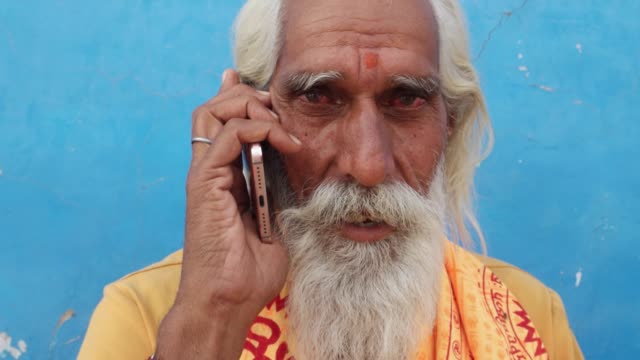 Hindu-Sadhu-Heiliger-Mann-mit-grauem-Bart-sitzt-und-spricht-ruhig-auf-seinem-Handy