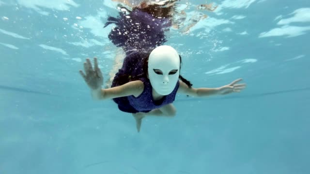 Ein-Kind-in-einer-weißen-Fee-Maske-schwimmt-und-stellt-unter-Wasser-in-einem-lila-Kleid,-sieht-in-die-Kamera-und-schwenkte-seine-Hände-auf-blauem-Grund.-Slow-Motion.
