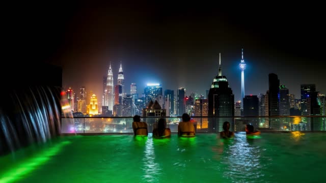Womans-swimming-to-the-edge-of-rooftop-swimming-pool-to-enjoy-the-night-cityscape-of-Kuala-Lumpur,-Malaysia-Timelapse-4K