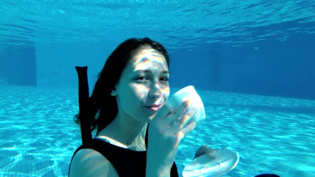 Beautiful-young-girl-sitting-underwater-at-the-bottom-of-the-pool-in-a-dress,-holding-a-white-Cup-and-saucer,-looking-at-the-camera-and-smiling-on-a-Sunny-day.-Slow-motion.-Zoom.-Close-up.
