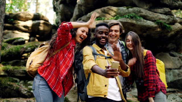 Glücklicher-afroamerikanischer-Kerl-macht-Videoanruf-mit-Smartphone-während-der-Wanderung-im-Wald-mit-Freunden,-junge-Menschen-sind-auf-der-Suche-am-Bildschirm,-winken-Hand-und-sprechen.
