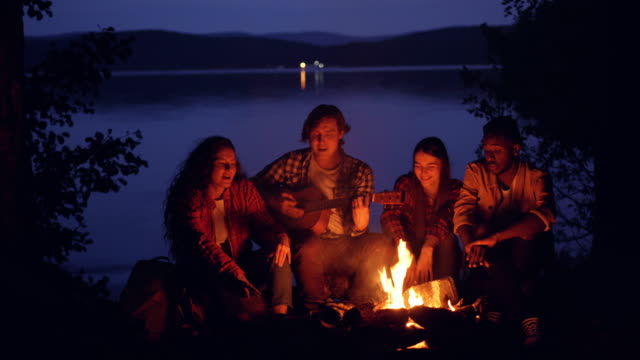 Happy-and-relaxed-guys-and-girls-are-singing-while-their-friend-is-playing-the-guitar-near-campfire-in-forest-near-lake-at-night.-Nature,-recreation-and-music-concept.