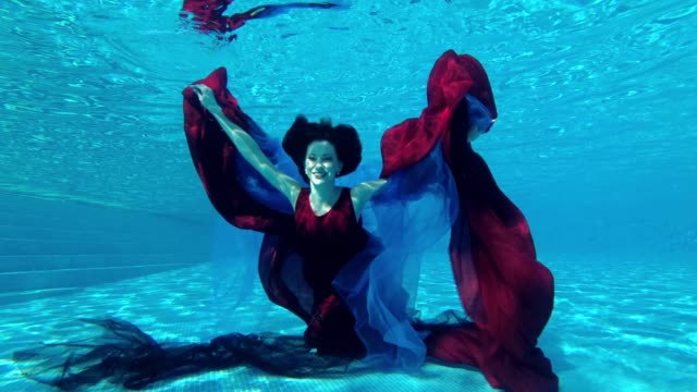Happy-girl-bride-in-red-dress-dives-under-the-water-to-the-bottom-of-the-pool-with-red-and-blue-cloth-in-her-hands.-She-looks-at-the-camera-and-smiles.-Slow-motion