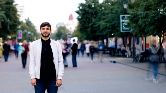 Time-lapse-of-good-looking-bearded-guy-looking-at-camera-and-smiling-standing-in-the-street-downtown-then-stepping-back-while-men-and-women-are-moving-near-him.