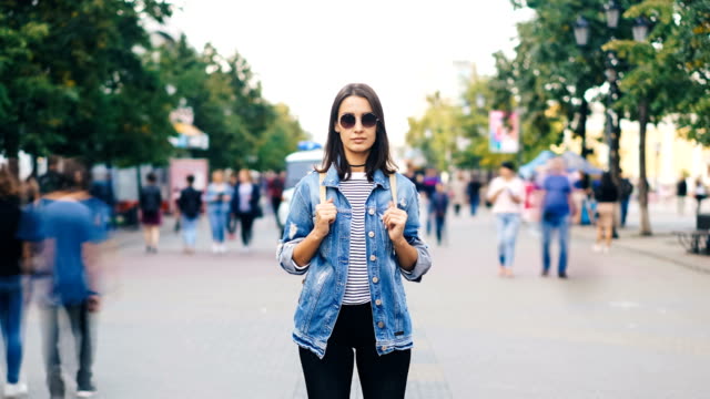 Zoom-out-time-lapse-of-confident-young-lady-in-sunglasses-looking-at-camera-standing-in-busy-pedestrian-street-in-flow-of-people.-Youth-and-society-concept.