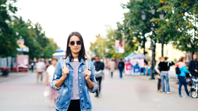 Time-lapse-portrait-of-confident-young-lady-in-sunglasses-standing-in-city-center-among-running-people-and-resting-looking-at-camera.-Time-and-youth-concept.