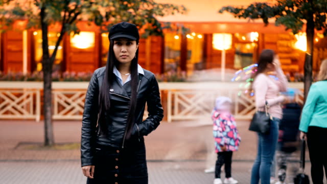 Time-lapse-portrait-of-tired-Asian-girl-standing-in-pedestrian-street-with-hand-in-pocket-and-looking-at-camera-then-leaving,-men-and-women-are-whizzing-around.