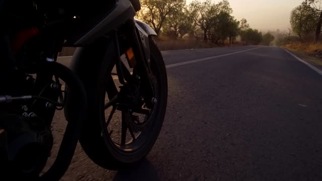 cinematic-shot-of-a-motorcycle-rider-in-a-sunset-time-crossing-the-road-in-the-forest,-seeing-some-many-parts-of-the-motorcycle-and-the-sky-and-the-trees,-camera-is-taking-the-wheel-crossing-the-road-between-the-fog