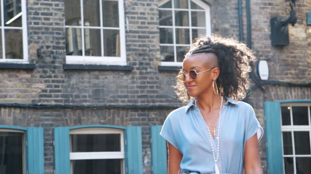 Trendy-young-black-woman-in-blue-dress-and-round-sunglasses-walking-on-the-street-past-camera,-backlit