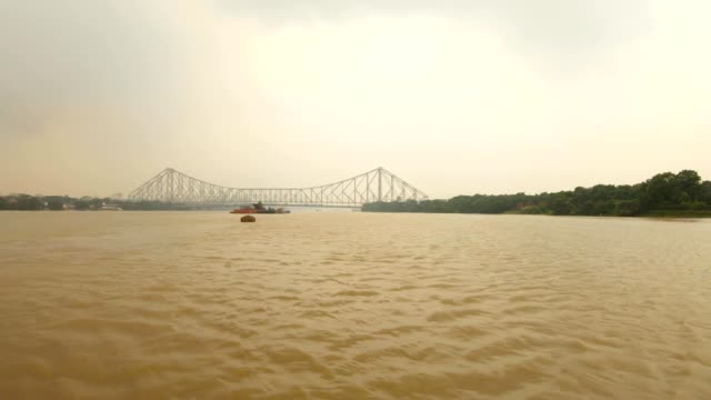 Howrah-bridge-full-flowing-river-Hoogli-barge-floats-buildings-and-trees-on-banks-cloudy-day