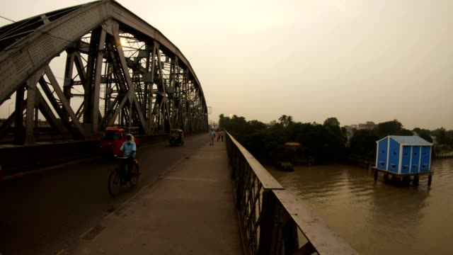Nivedita-Brücke-Dämmerung-blaues-Haus-auf-Stelzen-im-Fluss-Hooghly-Autos-Motorräder-und-Fußgänger-auf-der-Straße-Kolkata