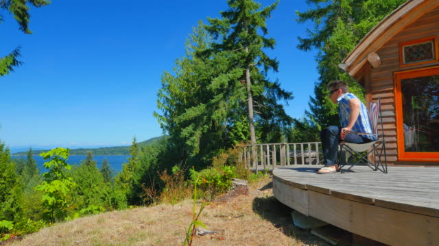 Video-Summer-Cabin-view-from-Bluff,-Man-Walks-and-Sits-in-Chair