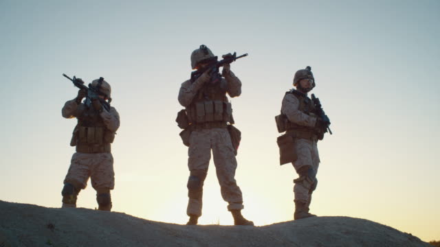 Truppe-von-drei-vollwaffenen-und-bewaffneten-Soldaten,-die-in-der-Wüstenumgebung-im-Sonnenuntergang-stehen.-Zeitlupe.