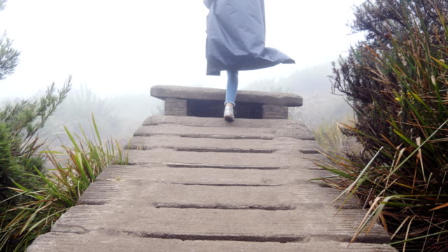 Young-female-tourist-at-raincoat-walking-upstairs-of-rocky-bridge-outdoor.-Woman-going-up-on-stone-stairs-during-vacation-travel-in-mountain.-Beautiful-nature-at-background.-Healthy-active-lifestyle
