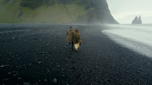 Luftaufnahme-des-Ehepaars-junge-Hipster,-die-auf-der-schwarzen-vulkanischen-Strand-in-der-Nähe-der-Troll-Zehen-in-Island-zurück
