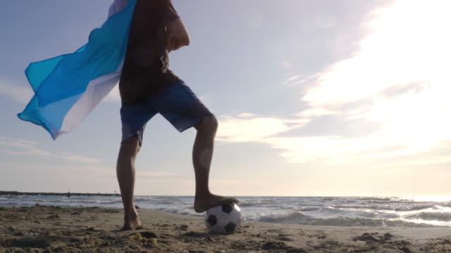 Young-man-wearing-argentinian-argentina-national-flag-as-super-hero-cape-stands-with-foot-on-football-on-the-sea-shore-sand-looking-at-the-ocean-at-the-beach-at-sunset-camera-steadycam-gimbal-revolving-around
