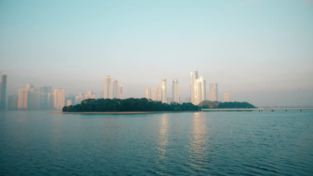 Urban-background,-morning-cityscape-with-skyscrapers,-blue-sky.-Buildings-reflecting-in-blue-sea.-Green-island-locked-shot.