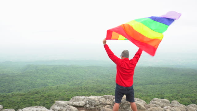 man-raise-rainbow-colour-LGBTI-flag-waving-in-hard-wind-on-mountain-top-viewpoint