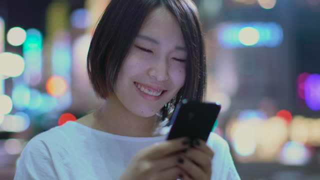 Porträt-von-die-attraktive-Japanerin-und-lässige-Kleidung-nutzt-Smartphone.-Im-Hintergrund-Plakate-Großstadt-Werbung-Lichter-leuchten-in-der-Nacht.