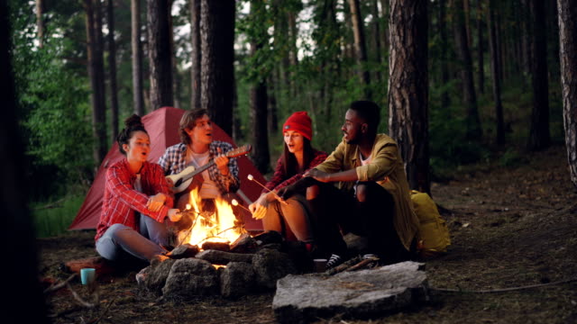 Relaxed-men-and-women-travelers-are-singing-songs-around-campfire-in-forest,-playing-the-guitar-and-roasting-marshmallow.-Green-trees-and-tent-is-visible.