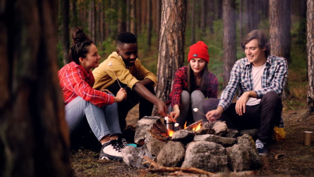 Hungry-travelers-are-cooking-marshmallow-on-fire-and-eating-it-from-sticks-during-conversation-around-campfire,-people-are-talking-and-laughing-enjoying-sweet-food.