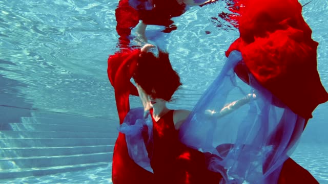 A-beautiful-girl-in-a-red-dress-with-red-hair-swims-and-poses-underwater-in-an-outdoor-pool-with-a-red-and-blue-cloth-in-her-hands.-She-looks-at-the-camera-and-smiles.-Vintage-processing.