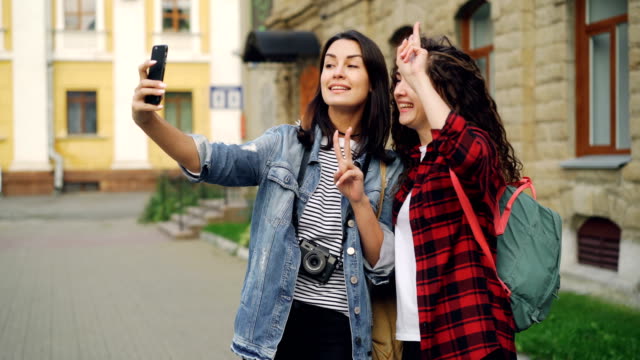 Cheerful-girls-foreign-travelers-are-taking-selfie-using-smartphone-standing-outdoors-and-posing-with-hand-gestures-showing-v-sign-and-heart-with-fingers-and-laughing.