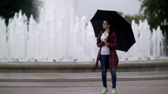 girl-with-an-umbrella-in-the-park-on-a-background-of-a-fountain,-looking-at-the-camera