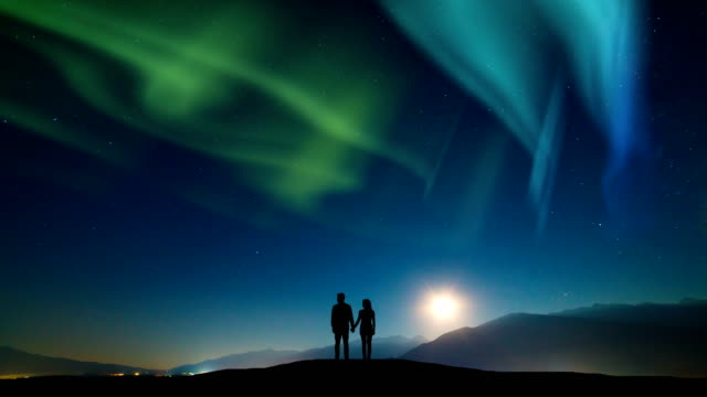 The-couple-standing-on-a-mountain-against-a-sky-with-northern-light.-time-lapse
