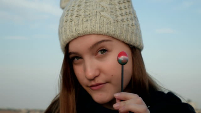 Pretty-teenage-girl-in-gray-knitted-hat-eating-lollipop-in-front-of-camera-and-smiling,-close-up-shot,-outdoor